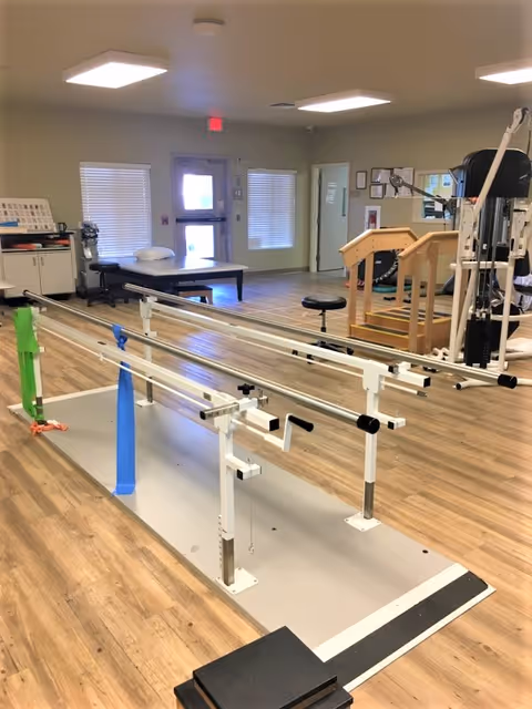 A physical therapy room with parallel bars for walking exercises, resistance bands attached to the bars, a padded treatment table, wooden stairs for rehabilitation, and various exercise equipment. The room has wood flooring, beige walls, and windows with blinds.