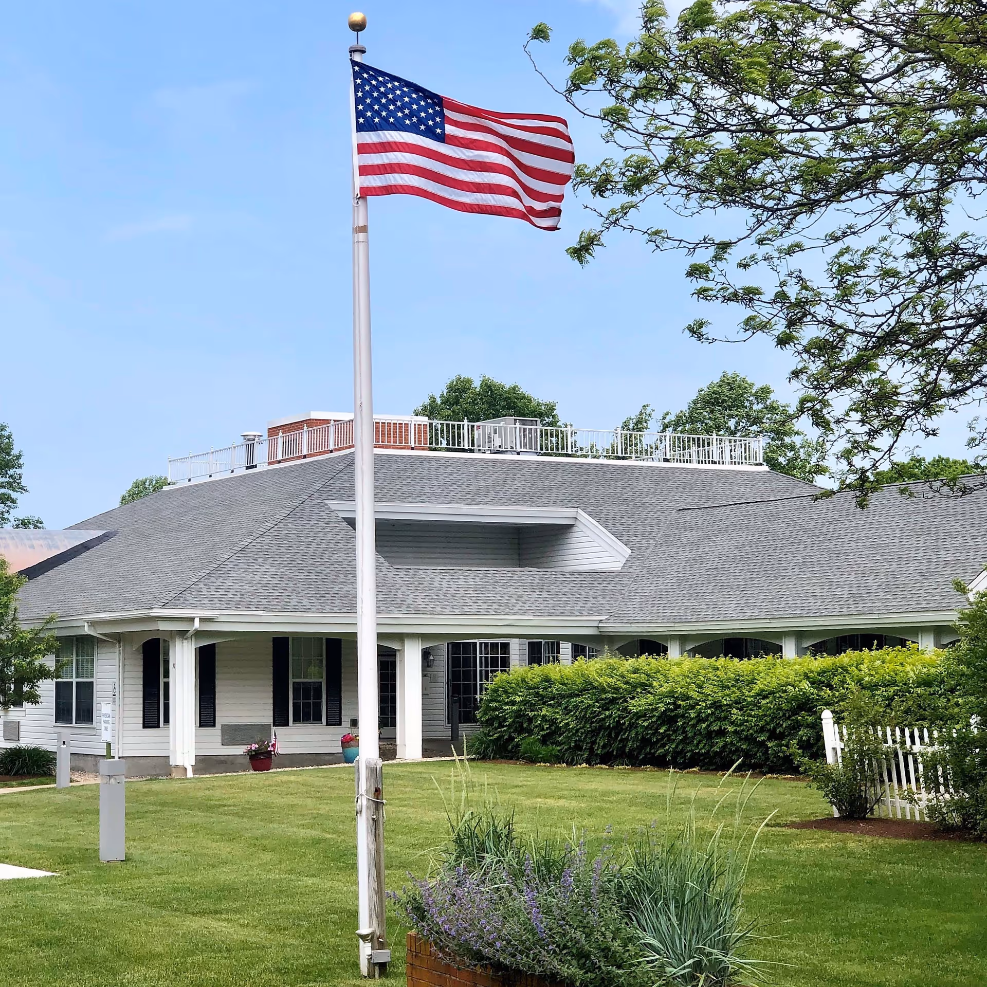 Exterior view of a single-story building with a gray shingled roof and white siding, surrounded by green bushes and a well-maintained lawn. An American flag on a flagpole is prominently displayed in the foreground, waving in the breeze under a clear blue sky.