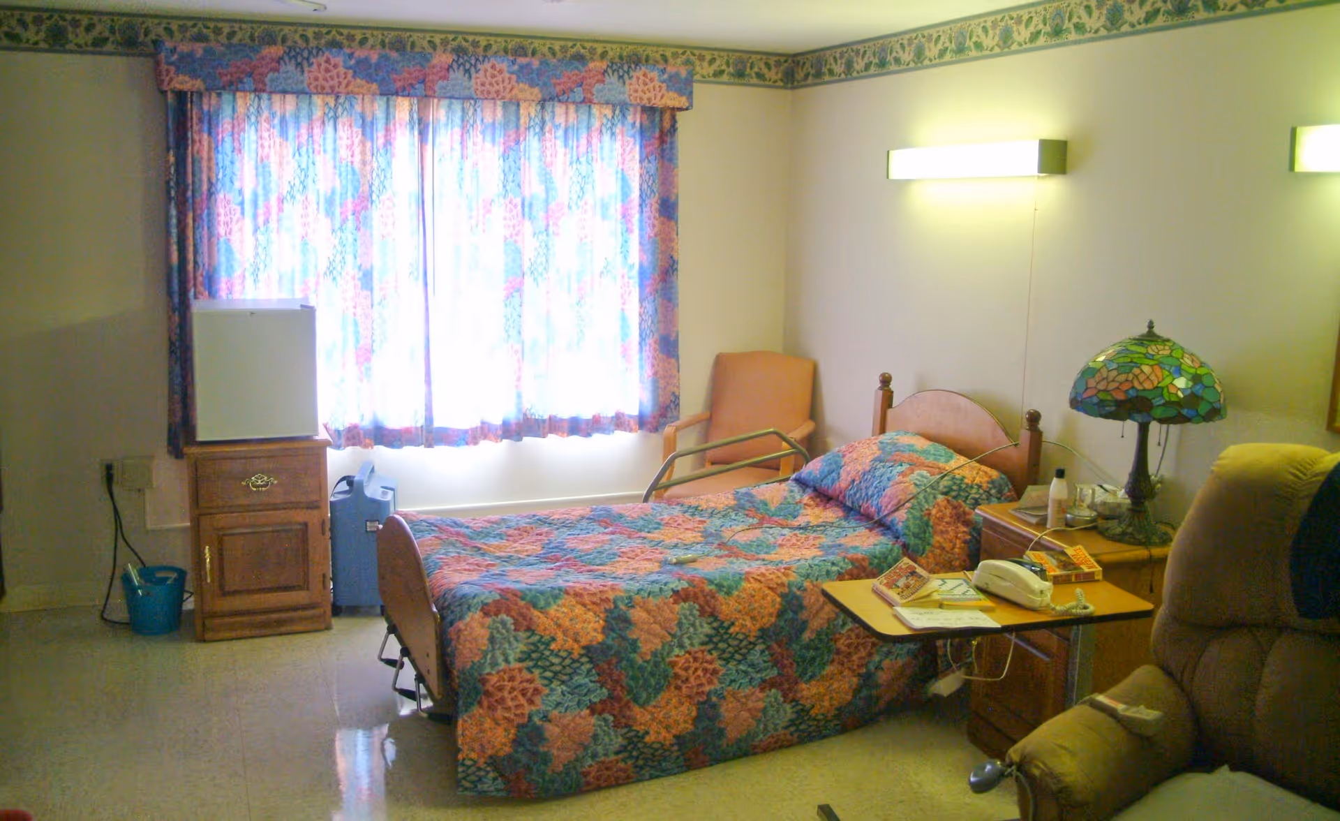 A furnished senior bedroom with a single bed covered in a colorful quilt, bedside tables, a Tiffany-style lamp, armchair, and a window with patterned curtains.