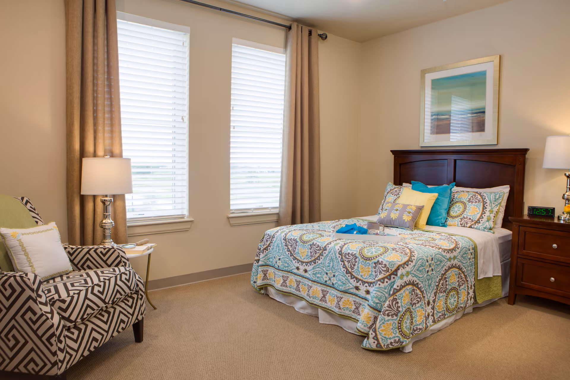 A cozy bedroom with a patterned bedspread in shades of blue, yellow, and brown on a wooden bed frame. There are multiple pillows on the bed, a wooden nightstand with a lamp and digital clock showing 12:57, and a patterned armchair with a white pillow. Two large windows with white blinds and beige curtains let in natural light. A framed abstract artwork hangs above the bed.