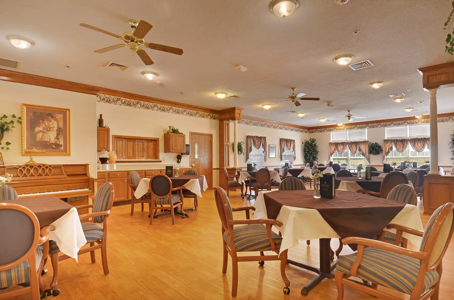 A spacious dining room in a senior living facility with multiple round tables covered with white and brown tablecloths, surrounded by wooden chairs with striped cushions. The room features wooden flooring, ceiling fans, large windows with valances, potted plants, a piano, and framed artwork on the walls.