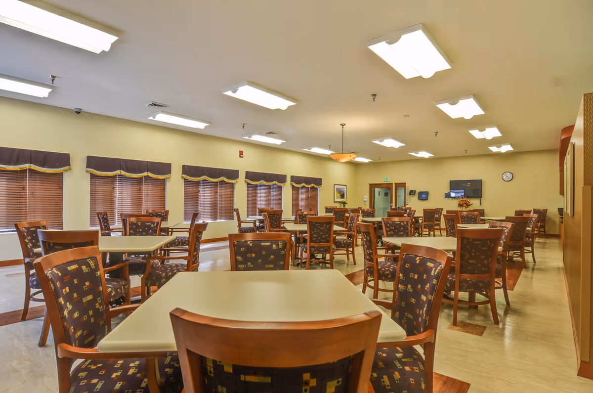 A spacious dining room with multiple square tables and wooden chairs featuring patterned upholstery. The room has beige walls, several windows with brown blinds and valances, and bright ceiling lights. A wall-mounted TV and a clock are visible on the far wall.