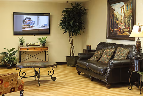 A cozy living room area with a dark leather sofa adorned with patterned cushions, a round metal coffee table with a glass top, a wooden side table with a lamp, a large framed painting on the wall, a flat-screen TV mounted on the wall, and several potted plants placed around the room on a wooden floor.