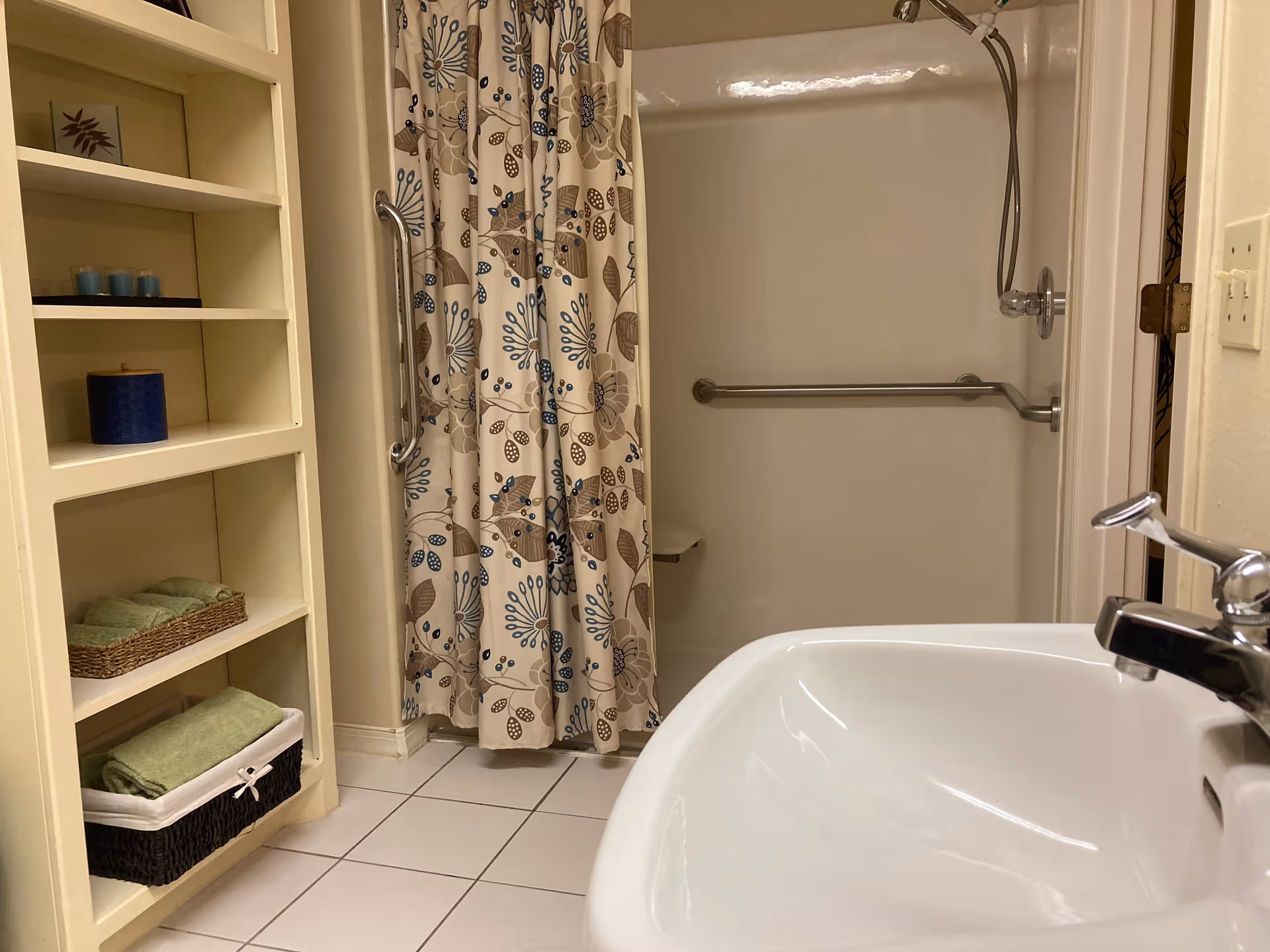 A bathroom with a white sink and chrome faucet in the foreground, a shower with a floral patterned curtain and grab bars, and a built-in shelving unit holding folded green towels, candles, and a small container.