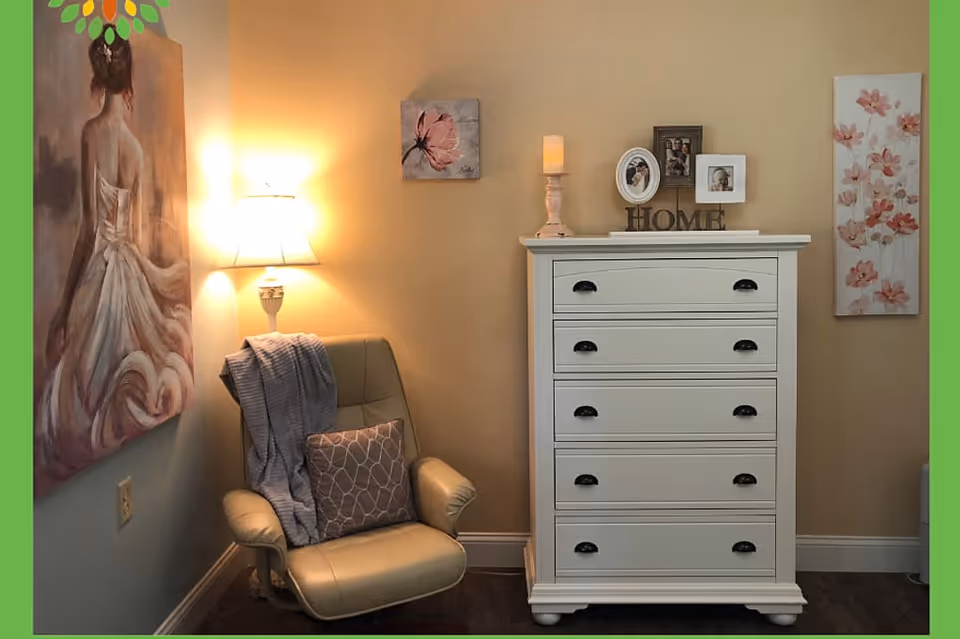 A cozy corner of a room featuring a beige leather armchair with a gray throw blanket and patterned pillow, a white chest of drawers with decorative items including framed photos, a candle, and a 'HOME' sign on top. The walls are adorned with floral paintings and a painting of a woman in a flowing dress. A wall lamp provides warm lighting.