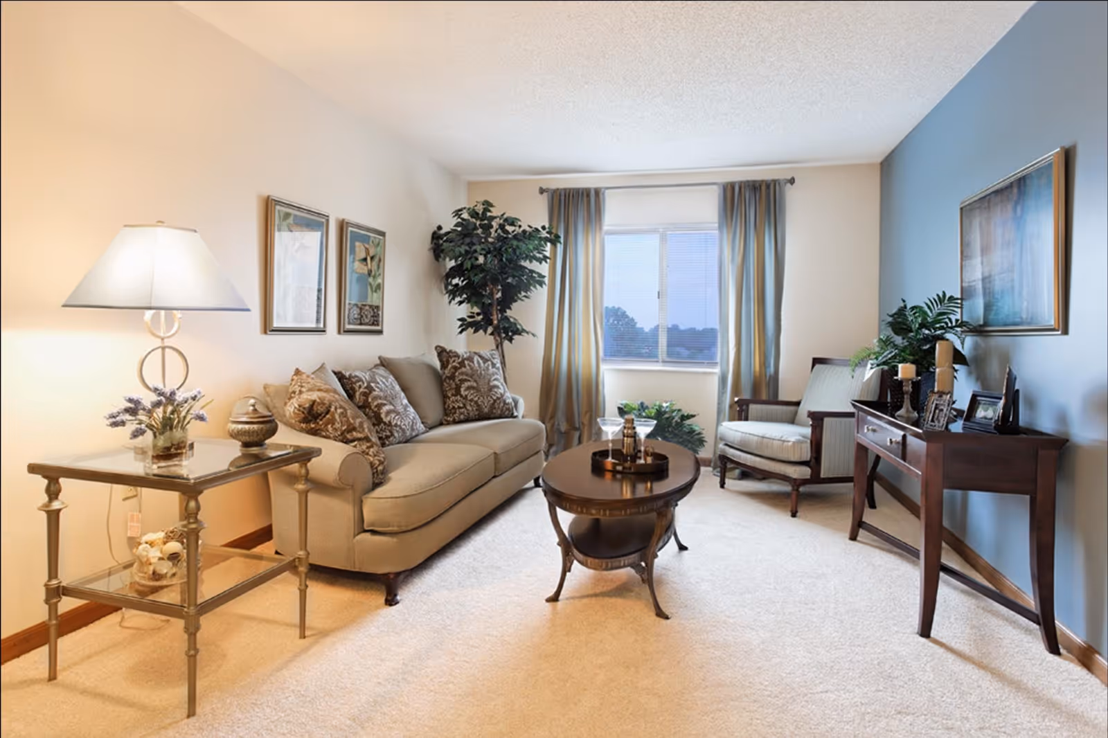 A cozy living room with a beige sofa adorned with patterned pillows, a glass side table with a lamp and decorative items, a wooden coffee table with a tray and glasses, a cushioned armchair, a wooden console table with candles and framed photos, a large window with curtains, and framed artwork on the walls.