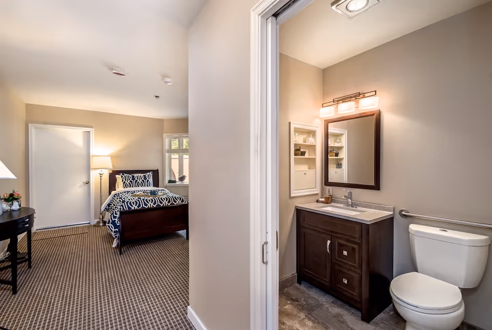 A furnished bedroom with a bed and nightstand visible alongside an open doorway revealing a small bathroom with a vanity and toilet.