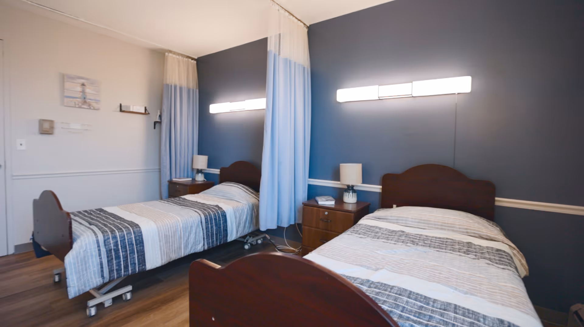 A room with two single beds separated by a blue privacy curtain. Each bed has a striped bedspread in shades of gray, white, and blue. There are wooden headboards and a small wooden nightstand between the beds with a lamp and a book. The walls are painted dark blue and white, and there is a bright horizontal light fixture above each bed.