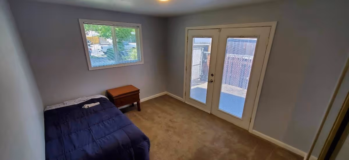 A small bedroom with a single bed covered in a dark blue comforter, a wooden nightstand next to the bed, a window with blinds on the left wall, and a set of glass double doors leading outside on the right wall. The room has beige carpet and light gray walls.
