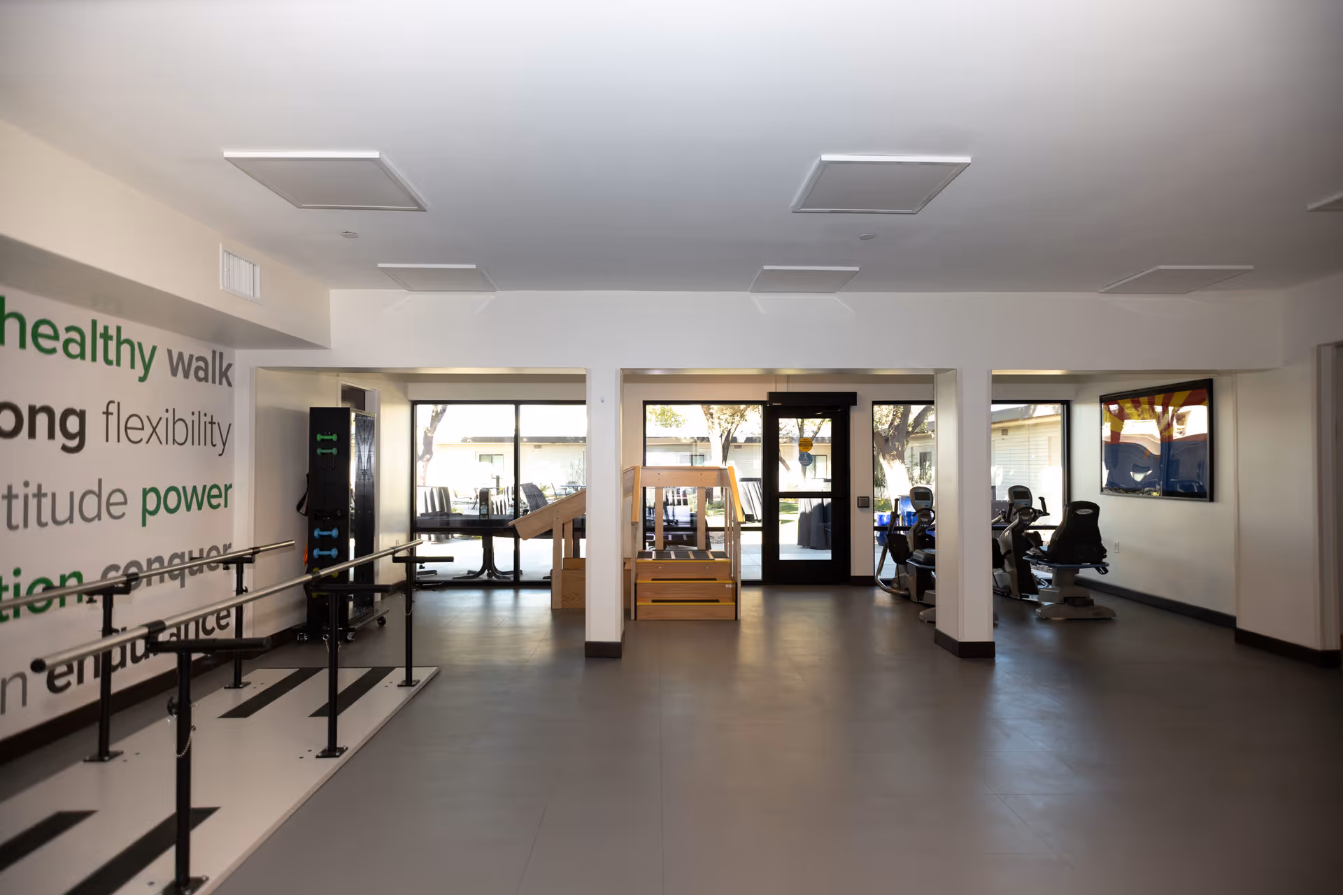 Interior view of a rehabilitation or fitness room with parallel bars for walking exercises on the left, exercise equipment on the right, and a wooden staircase in the center. Large windows and a glass door provide natural light and a view of the outside.
