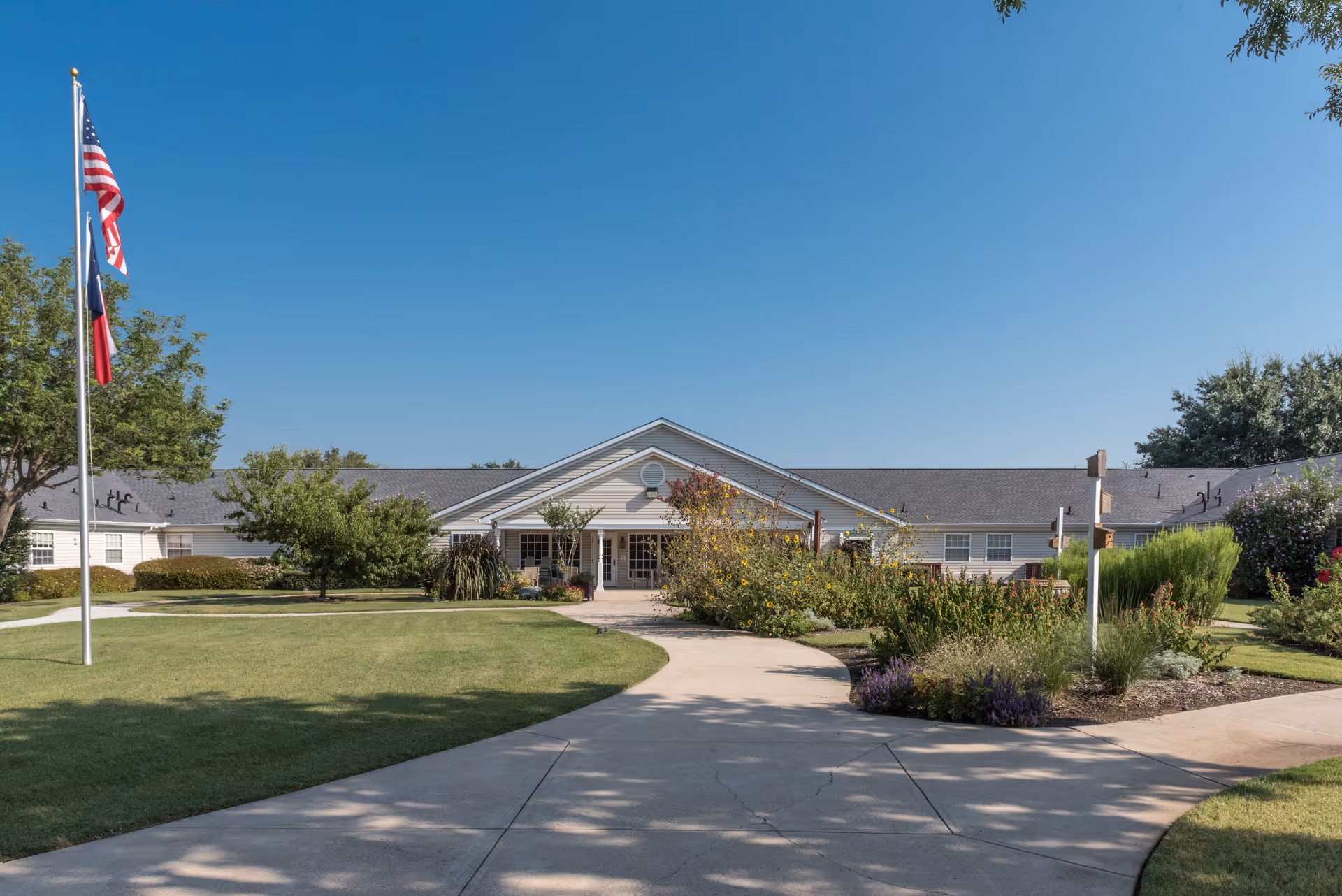 Front exterior view of Arden Courts Of Richardson building with a paved walkway leading to the entrance, surrounded by green lawns, trees, and flower beds under a clear blue sky. Two flagpoles with the American and Texas flags are visible on the left side.