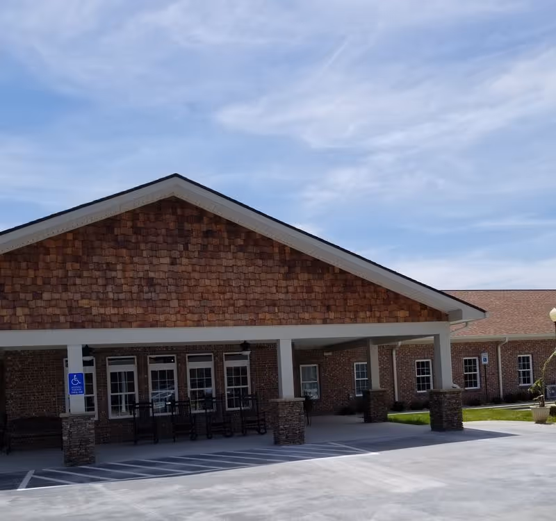 Front entrance of a brick senior living building with a covered portico, rocking chairs, and marked handicapped parking under a blue sky.