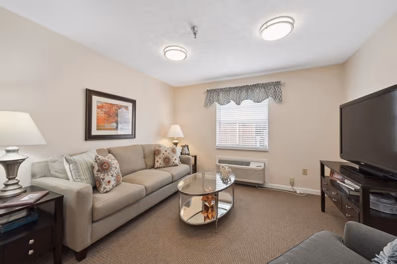 A cozy living room with a beige sofa adorned with patterned pillows, a glass oval coffee table with decorative items, a flat-screen TV on a dark wooden stand, a window with blinds and a valance, two table lamps, and a framed picture of autumn trees on the wall.