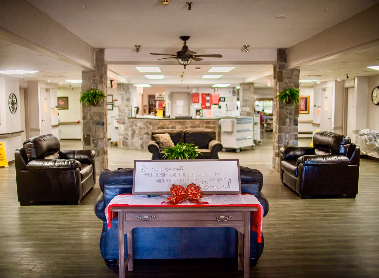 A cozy nursing home common area with dark leather armchairs and a loveseat arranged around a small wooden table. The table has a red cloth and a decorative sign that reads 'Be our guest whether you come in to visit, or just to rest when you enter our home may you be blessed.' The room features stone pillars with hanging plants and a ceiling fan, with a reception desk visible in the background.