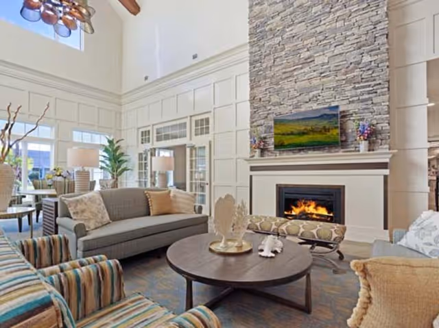 A spacious and well-lit living room with high ceilings, featuring a stone fireplace with a TV mounted above it. The room has comfortable seating including a gray sofa, striped armchairs, and a cushioned bench. A round wooden coffee table with decorative items sits in the center. Large windows and glass doors allow natural light to fill the space, and there are several lamps and plants adding to the cozy atmosphere.