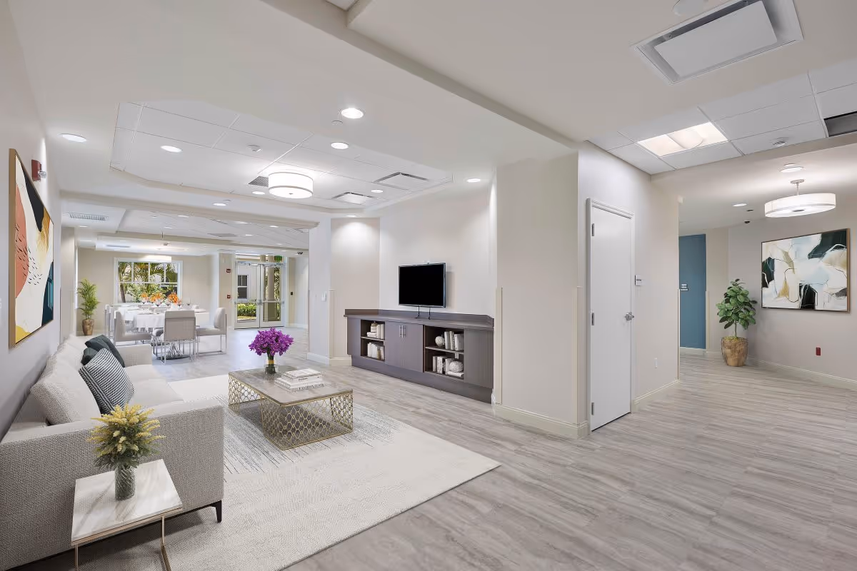 A modern senior living facility interior featuring a comfortable living room with a light gray sofa, a coffee table with a vase of purple flowers, and a wall-mounted TV above a cabinet with books. In the background, there is a dining area with a table set for a meal and large windows letting in natural light. The space has light-colored walls, wood-look flooring, and contemporary ceiling lights.