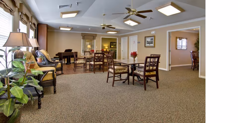 Communal lounge with dining tables and chairs, upholstered armchairs, plants, and ceiling fans.