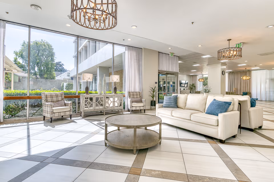 Bright and spacious senior living facility lounge area with large floor-to-ceiling windows letting in natural light. The room features a cream-colored sofa with blue pillows, two plaid armchairs, a round wooden coffee table, and a mirrored console table with two lamps. The floor has a white and gray tile pattern, and there are modern chandeliers hanging from the ceiling. Outside the windows, greenery and trees are visible.