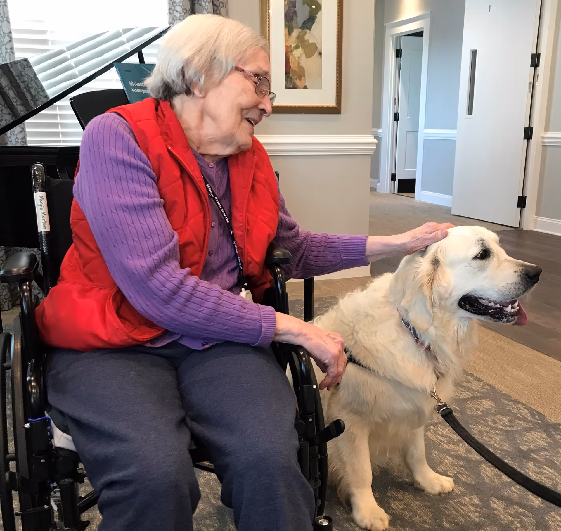 An elderly woman in a wheelchair wearing a red vest and purple sweater is smiling and petting a large white dog sitting beside her in a well-lit room with light-colored walls and carpeted floor.