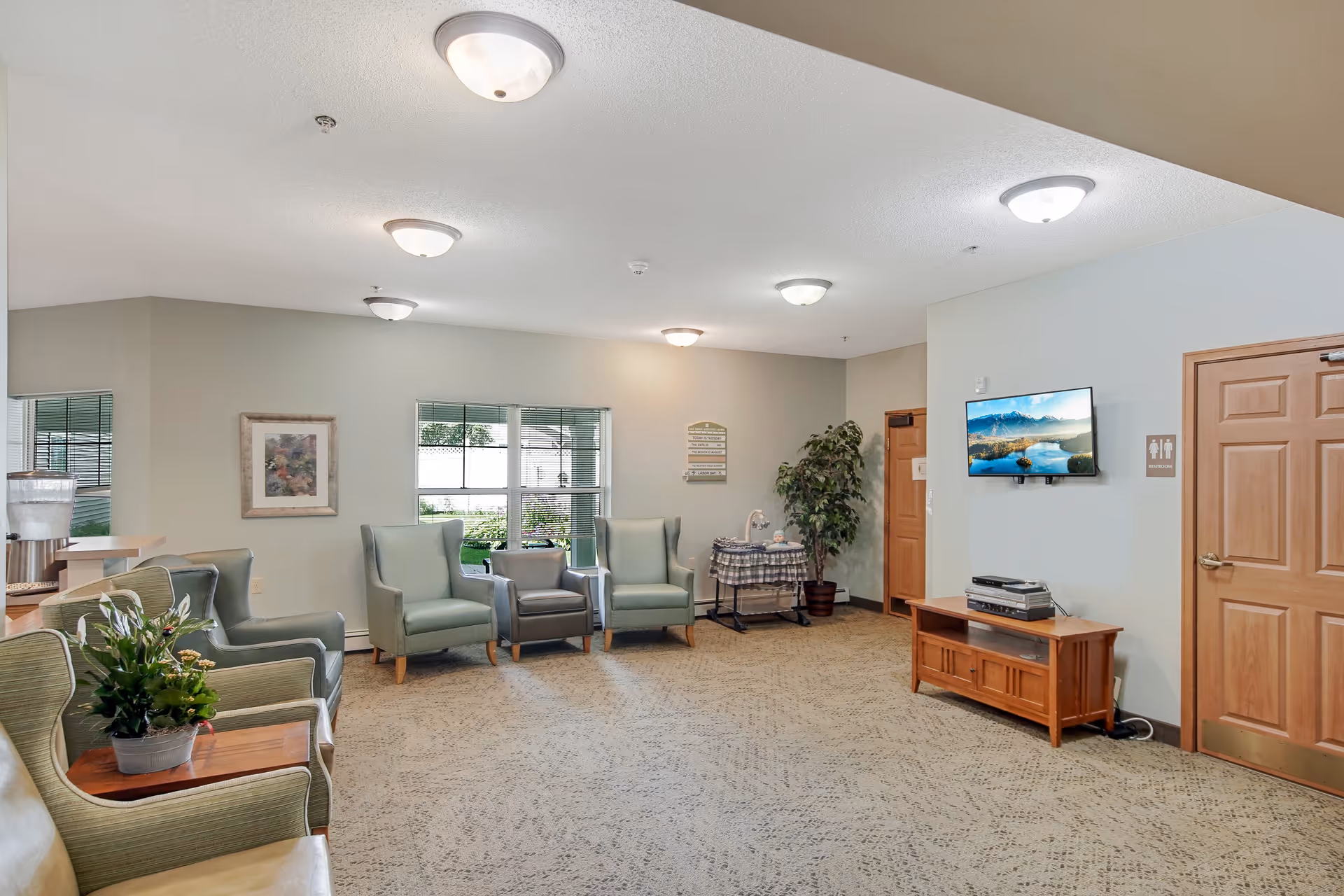 A spacious assisted living facility common area with several armchairs arranged along the walls, a small table with a plant, a water dispenser, a TV mounted on the wall displaying a scenic landscape, and two wooden doors, one marked with restroom signs. The room has beige walls, carpeted flooring, and ceiling lights.