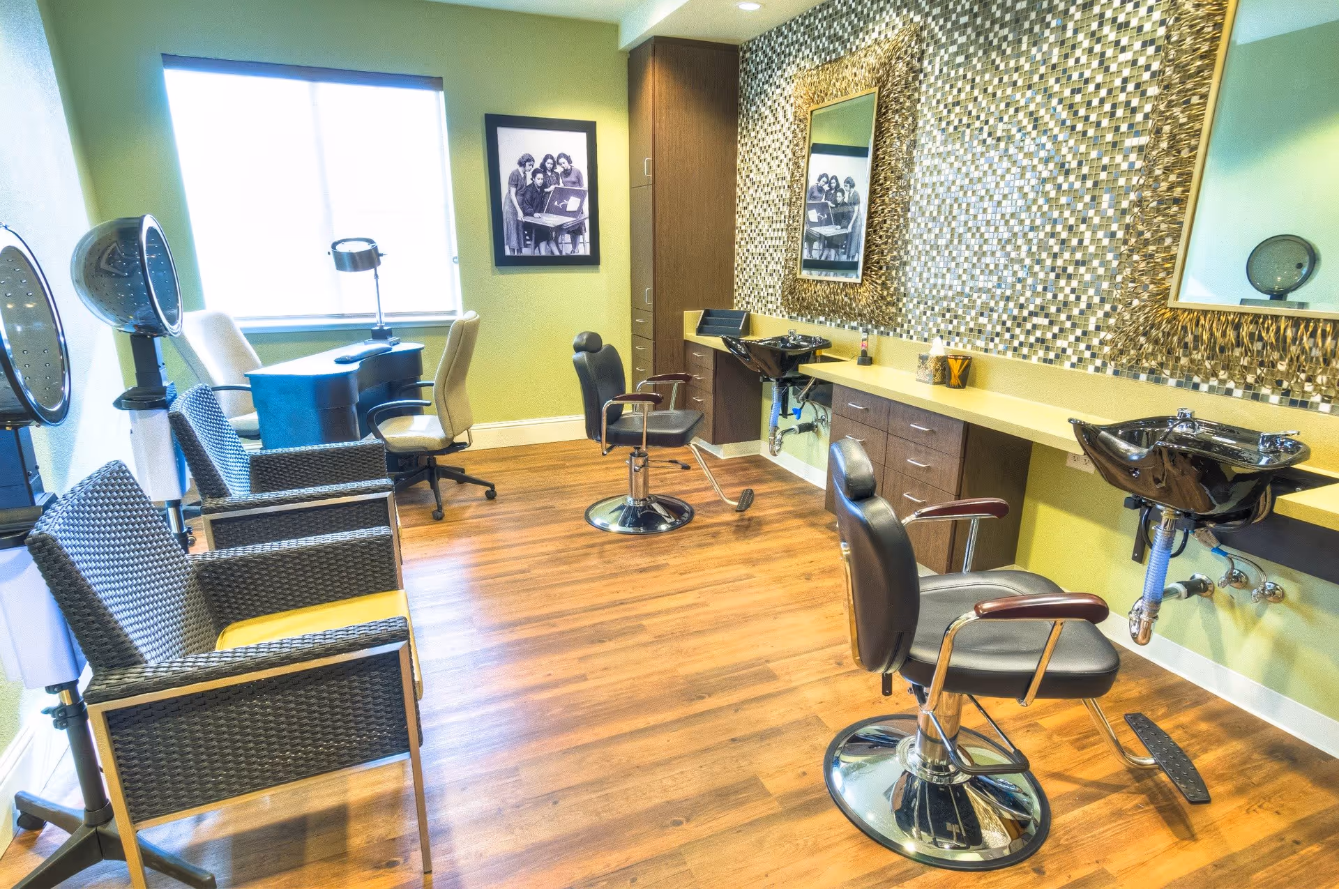 Interior of a salon room with wooden flooring, two black salon chairs in front of black wash basins, two wicker chairs, a desk with two office chairs near a window, and a wall decorated with mosaic tiles and framed black and white photos.