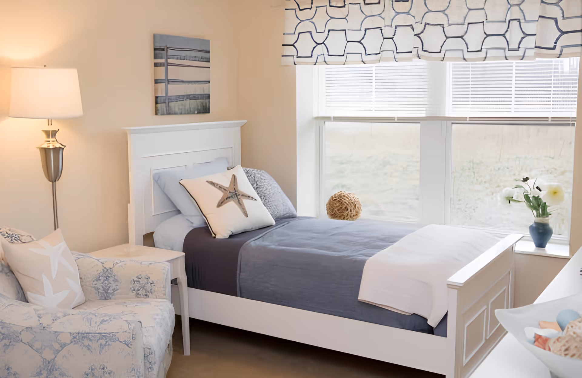 A cozy bedroom with a white bed featuring blue and white bedding, a starfish decorative pillow, a cushioned armchair with a matching starfish pillow, a floor lamp, a small side table, a window with patterned blinds, and a vase with flowers on the windowsill.