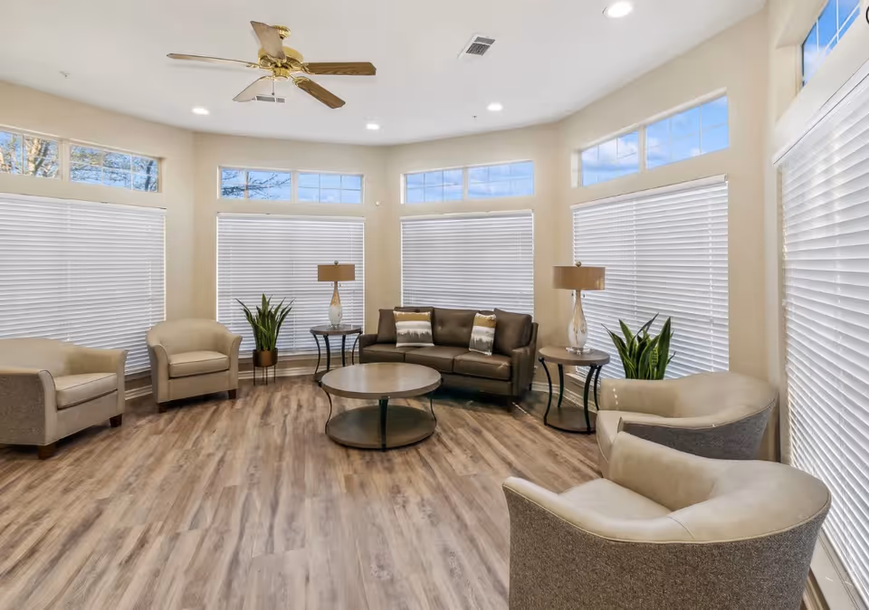 Spacious bright common living area with multiple armchairs, a sofa around a round coffee table, lamps, plants, and large windows with blinds.
