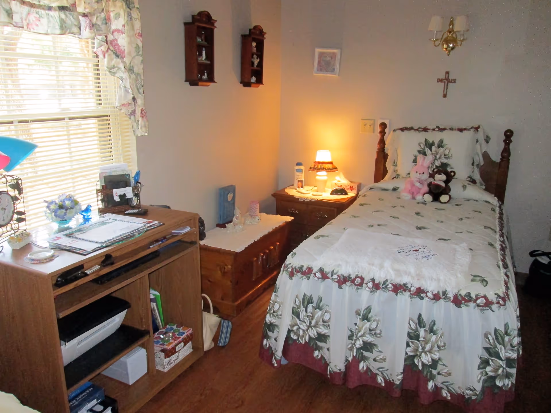 A cozy bedroom with a single bed covered in floral bedding and two stuffed animals placed on the pillow. Next to the bed is a wooden nightstand with a lamp, lotion, and other small items. A wooden chest with decorative items on top is against the wall. A wooden shelving unit with various items and a printer is near a window with floral curtains. The walls have small shelves with figurines, a framed picture, and a cross.