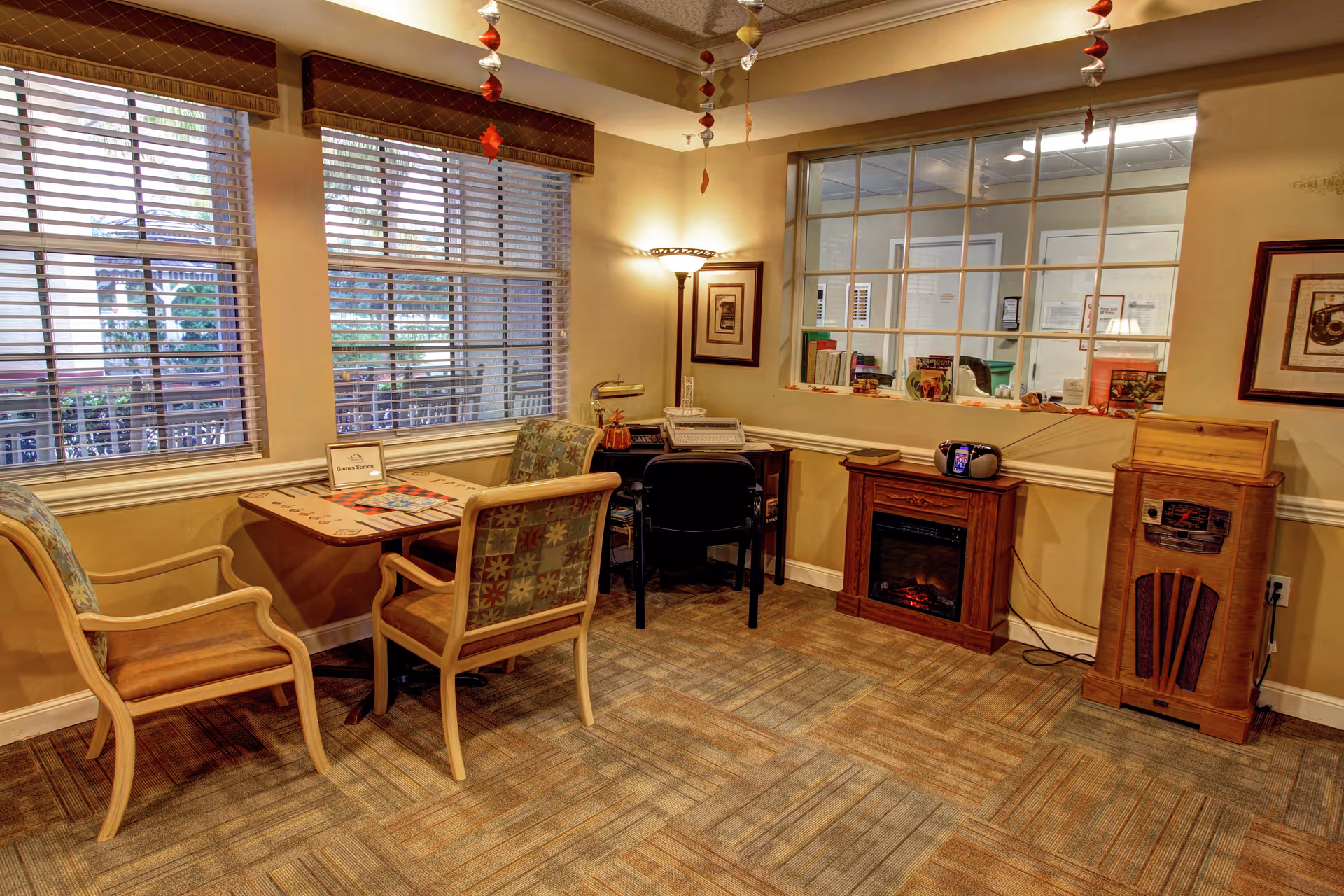A cozy interior room with a small table and three chairs near two large windows with blinds. There is a desk with a chair, a lamp, and a printer. A small electric fireplace and a vintage-style radio are also visible. The room has warm lighting and beige walls with framed artwork.