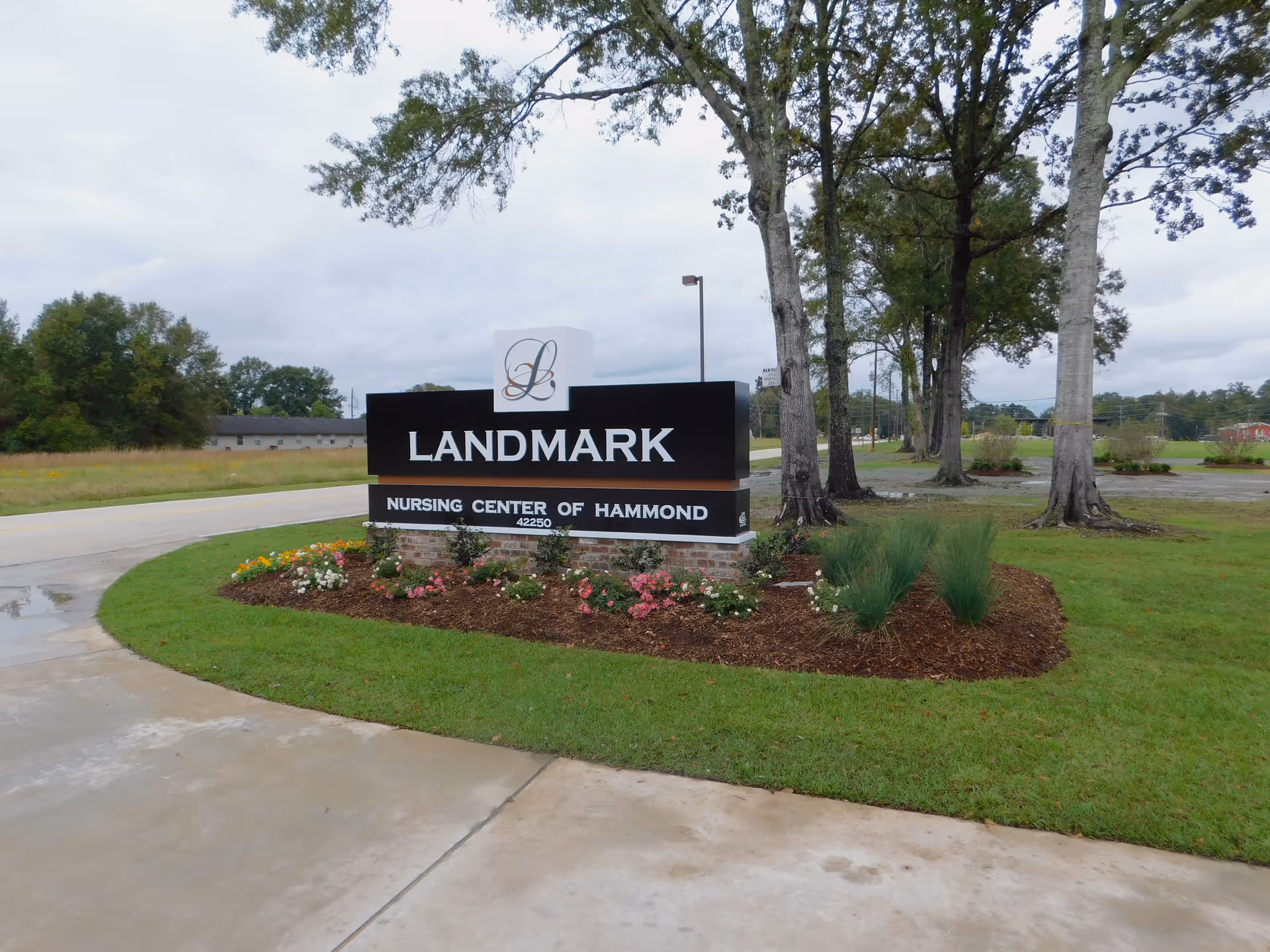 Outdoor view of the Landmark Nursing Center of Hammond sign surrounded by a landscaped flower bed with grass and trees in the background under a cloudy sky.