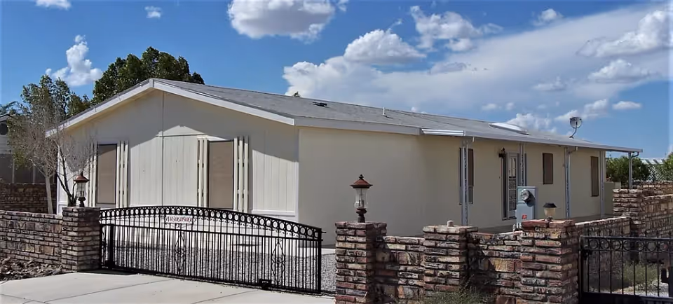 Single-story beige building with a sloped roof behind a black metal gate and brick fence under a partly cloudy blue sky.