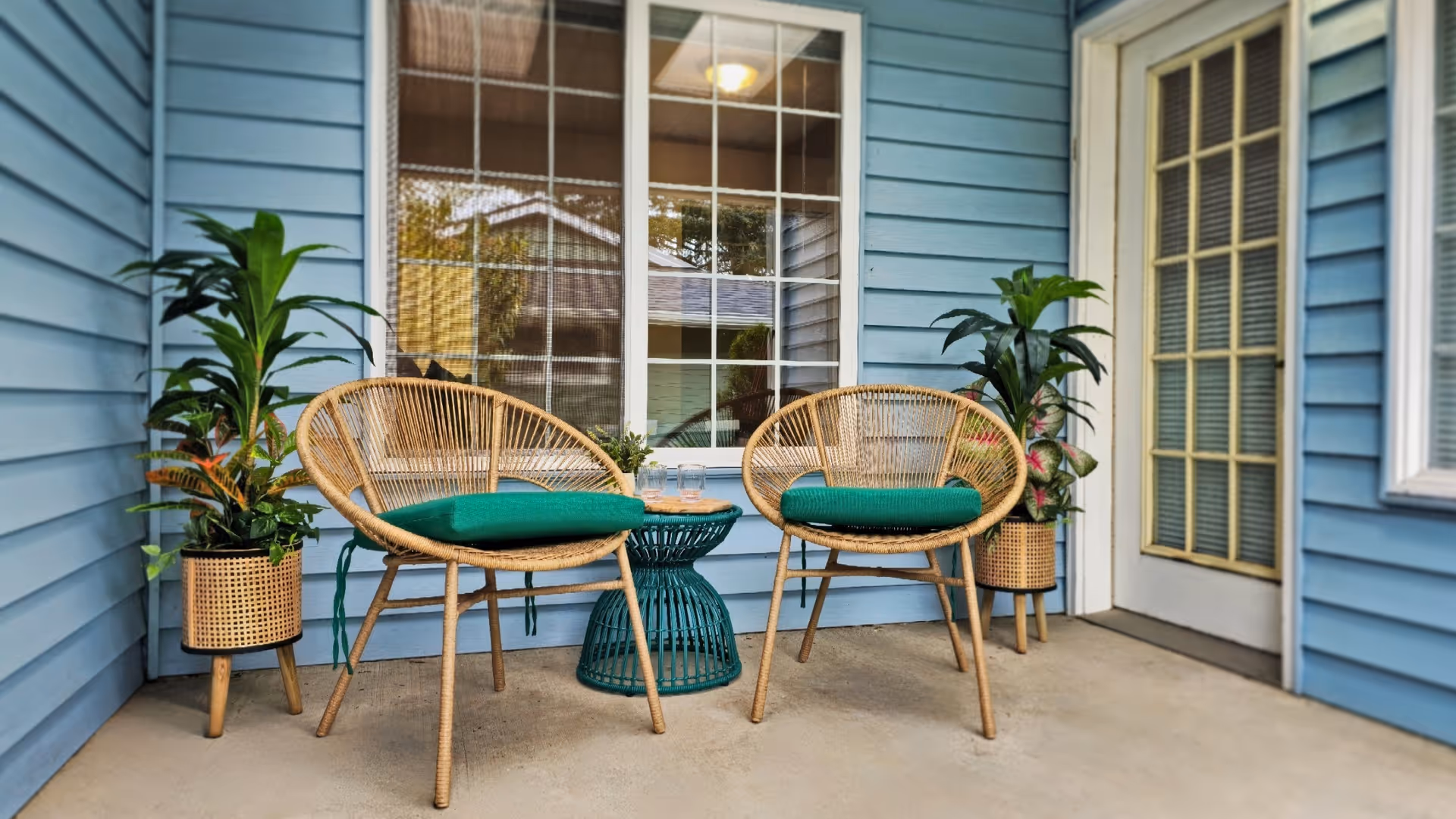 Two wicker chairs with green cushions and a small round table on a blue-painted porch beside a window and door.