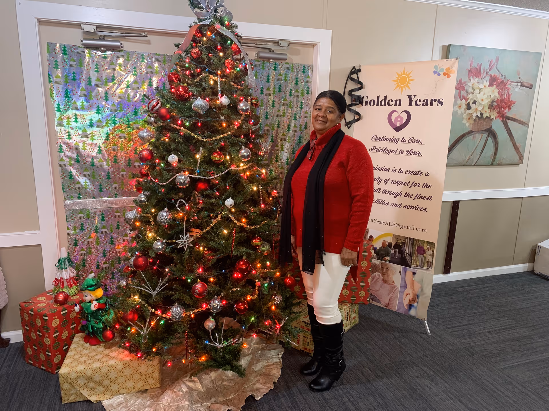 A woman dressed in a red sweater, white pants, black boots, and a black scarf stands next to a decorated Christmas tree with red and silver ornaments and lights. Wrapped presents and a small elf doll are placed under the tree. Behind her is a banner for Golden Years Assisted Living Facility with text about their mission, and a wall painting of a bicycle with flowers.