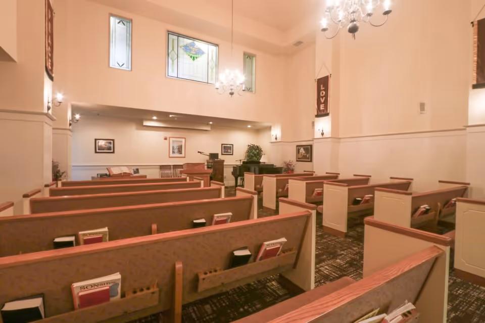 Interior view of a chapel or small worship room with rows of wooden pews, hymnals, and Bibles. The room has high ceilings with chandeliers, stained glass windows, wall sconces, and banners with the word 'LOVE'. There is a piano and a podium at the front of the room.