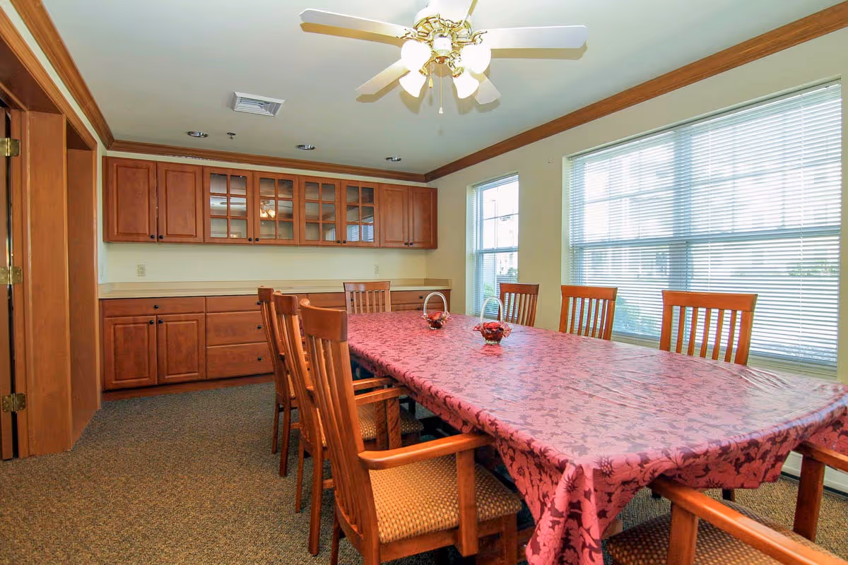 A dining room with a long rectangular table covered with a red patterned tablecloth, surrounded by wooden chairs with cushioned seats. The room has large windows with blinds allowing natural light to enter. There are wooden cabinets with glass doors mounted on the wall and a ceiling fan with lights above the table.