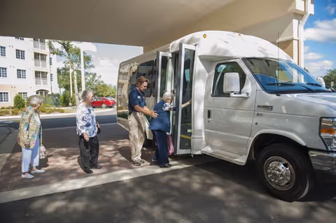 A group of elderly women boarding a white shuttle bus outside a building, assisted by a staff member under a covered driveway on a sunny day.