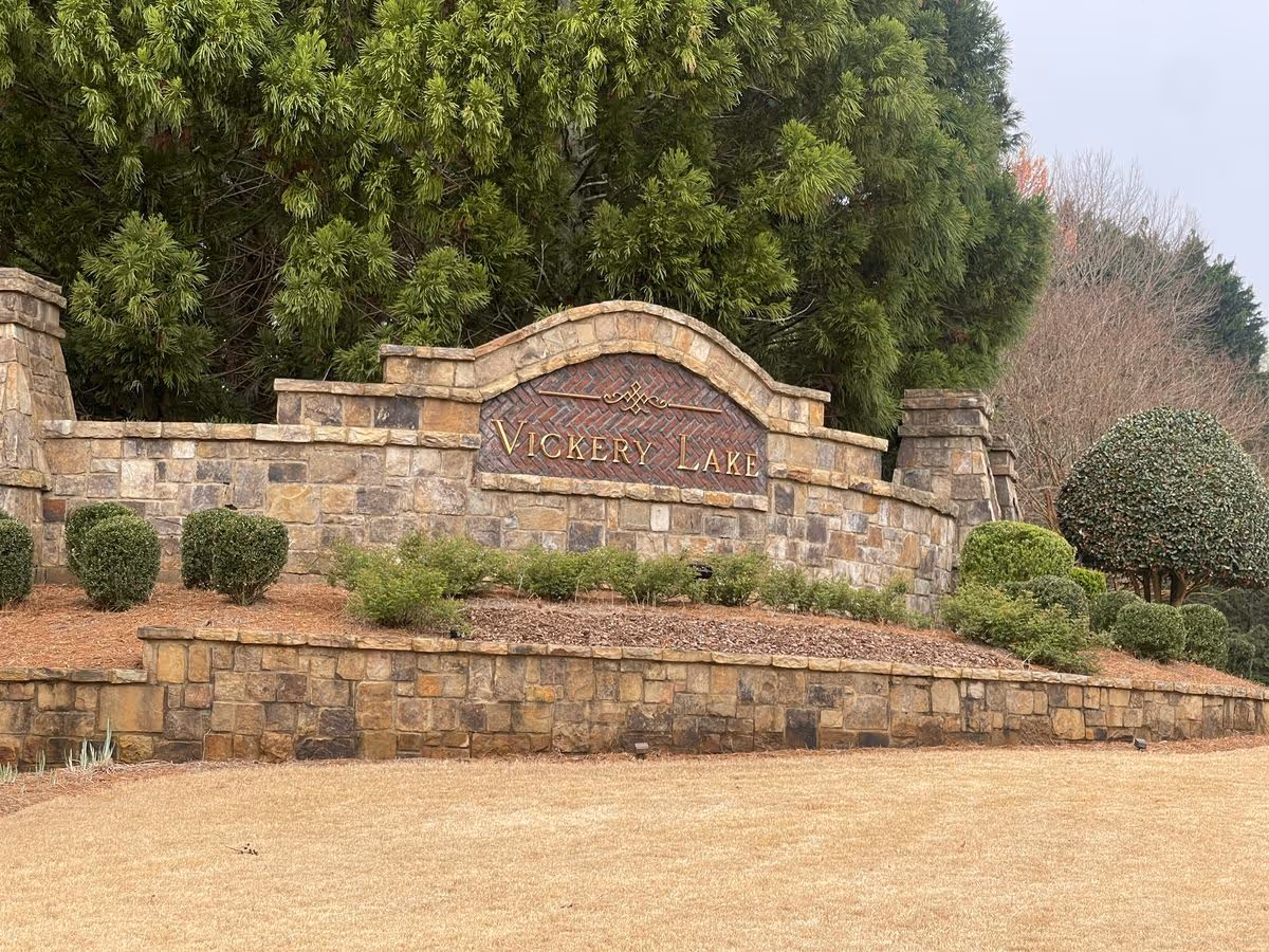 Stone entrance sign reading "Vickery Lake" set into a landscaped stone wall with shrubs and trees behind it.