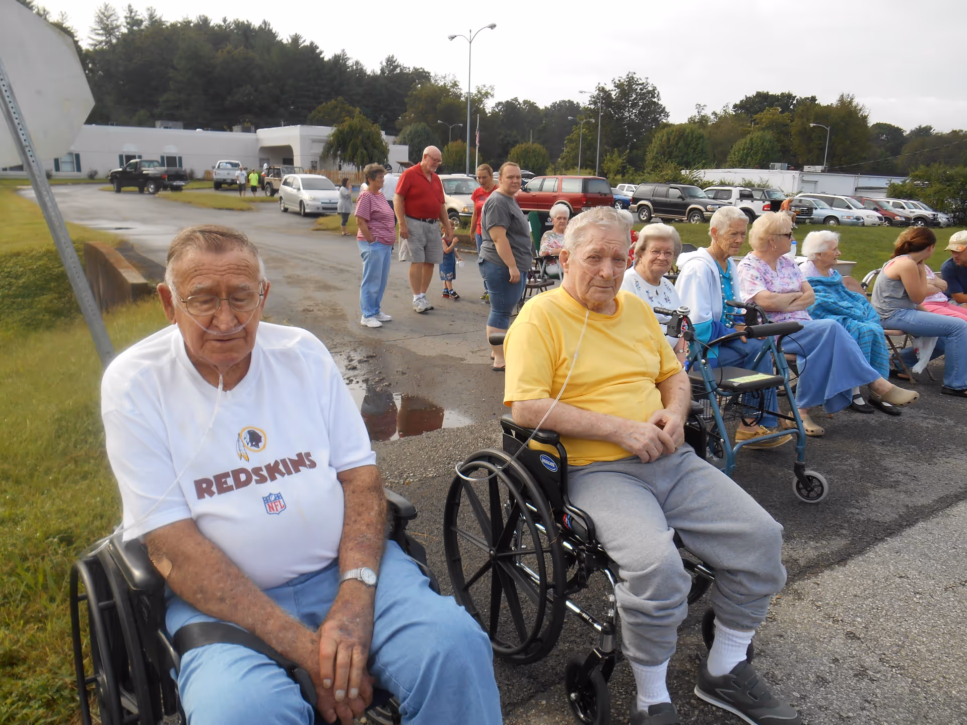 A group of elderly people, some in wheelchairs and others seated or standing, gathered outdoors on a paved area near a senior living facility. Two elderly men in the foreground are using oxygen tubes. Several other people, including caregivers, are standing or sitting in the background with cars and trees visible behind them.