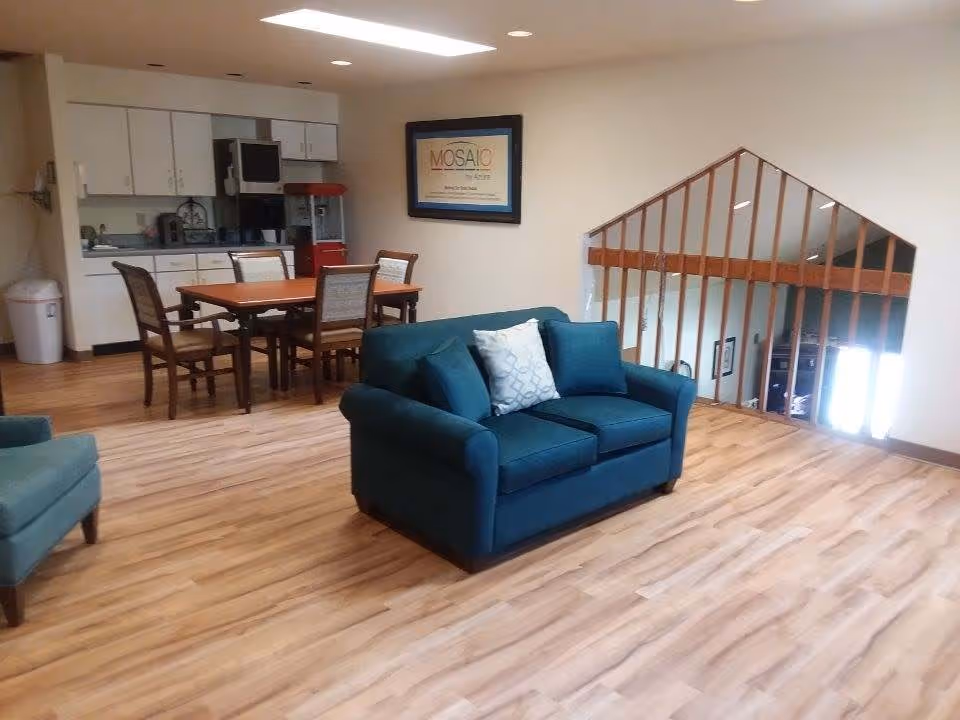 Interior view of a senior living facility common area featuring a teal loveseat with pillows, a dining table with four chairs, a kitchenette with white cabinets, and a popcorn machine. The room has wood flooring and a railing overlooking a lower level.