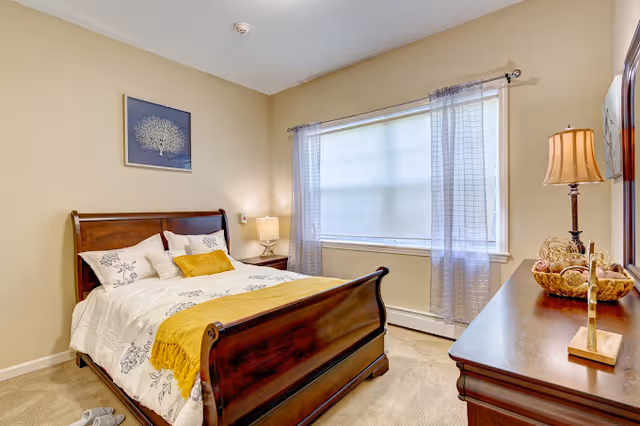 A cozy bedroom with a wooden sleigh bed dressed in white and yellow bedding. There is a nightstand with a lamp beside the bed, a large window with sheer curtains, and a wooden dresser with a lamp and decorative items. A framed artwork hangs on the beige wall above the bed.