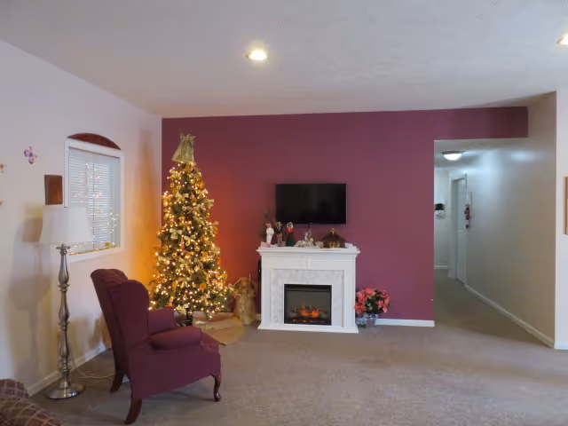 A cozy living room decorated for Christmas with a lit Christmas tree, a white electric fireplace, a wall-mounted TV, a burgundy armchair, a floor lamp, and holiday decorations including poinsettias and figurines. The walls are painted beige and dark red, and there is a hallway visible to the right.