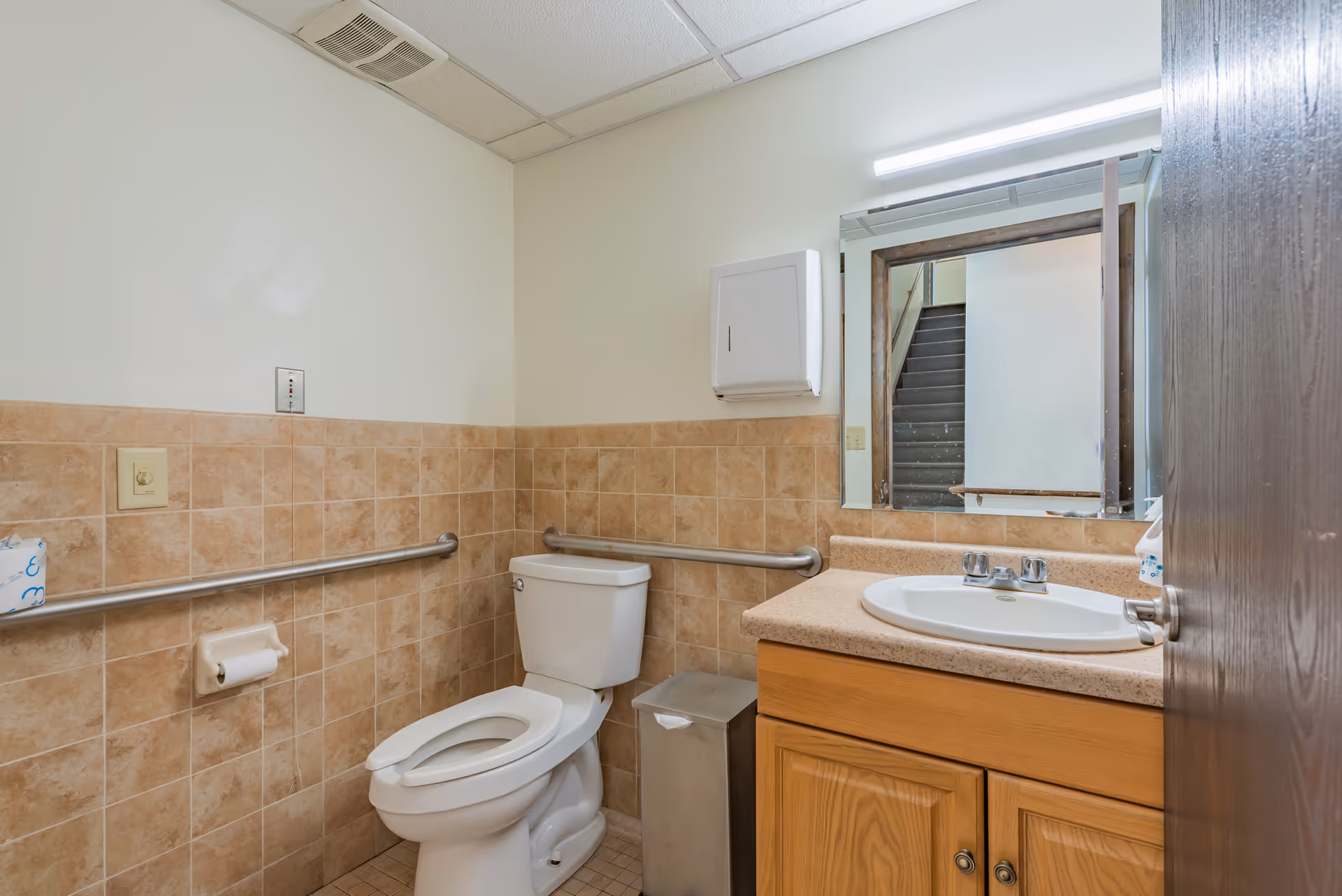 A clean bathroom with beige tiled walls and floor. It features a white toilet with a grab bar on the wall beside it, a wooden vanity with a beige countertop and a white sink, a large mirror above the sink, a wall-mounted paper towel dispenser, and a metal trash can next to the vanity. The ceiling has a vent and fluorescent lighting.