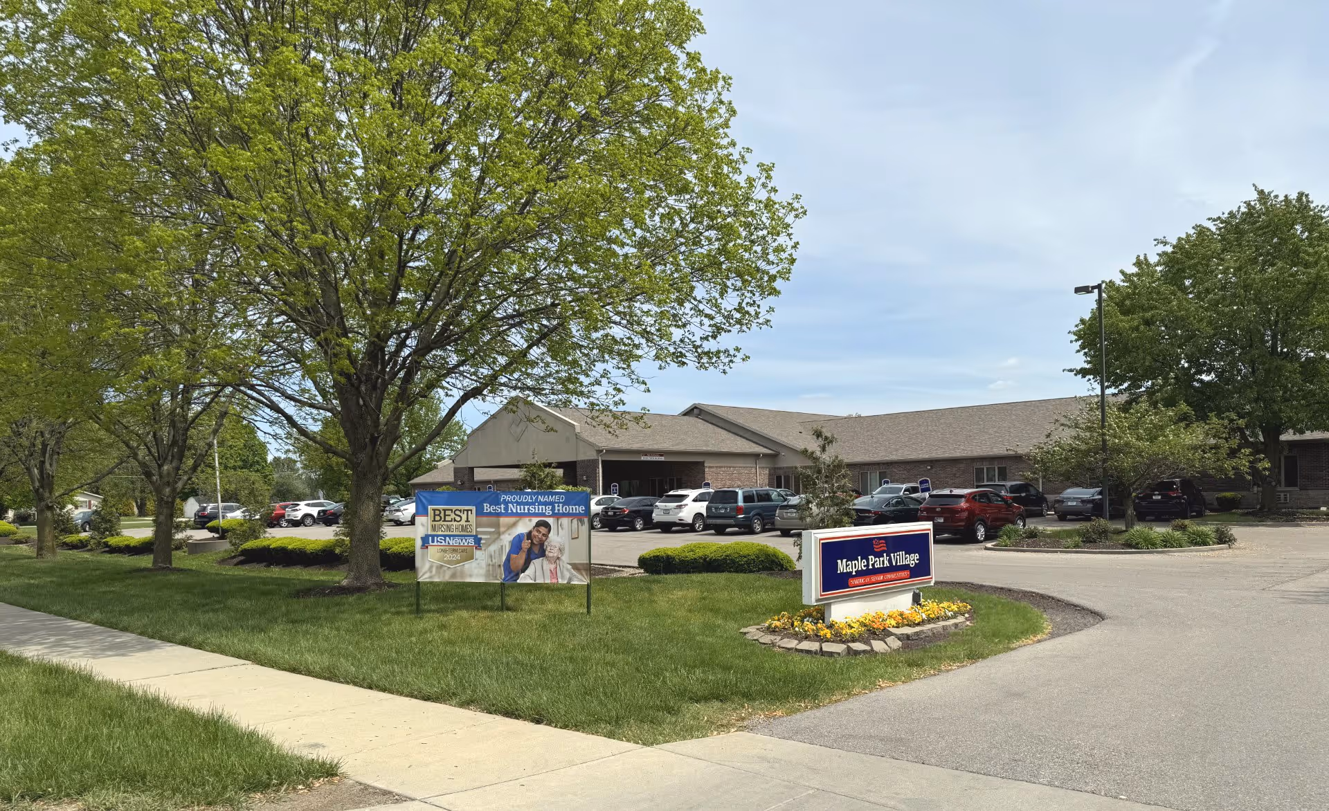 Exterior view of Maple Park Village senior living facility with a parking lot, green trees, and two signs on the lawn. One sign reads 'Maple Park Village' and the other sign highlights an award for Best Nursing Home by U.S. News.
