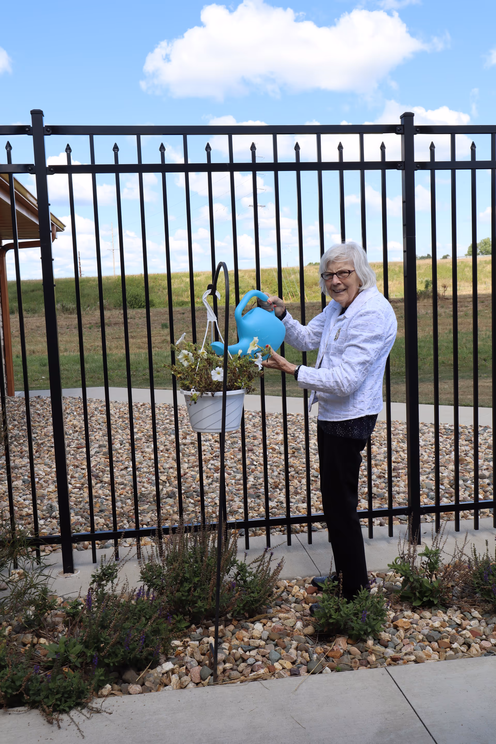 An elderly woman outdoors watering a hanging planter with a blue watering can in front of a metal fence.
