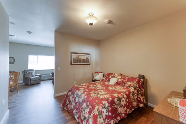 A cozy bedroom with a floral red bedspread on a double bed, a wooden nightstand with a clock and a basket, a framed picture on the beige wall, and a view into a living area with a window, armchair, and dining table with chairs.