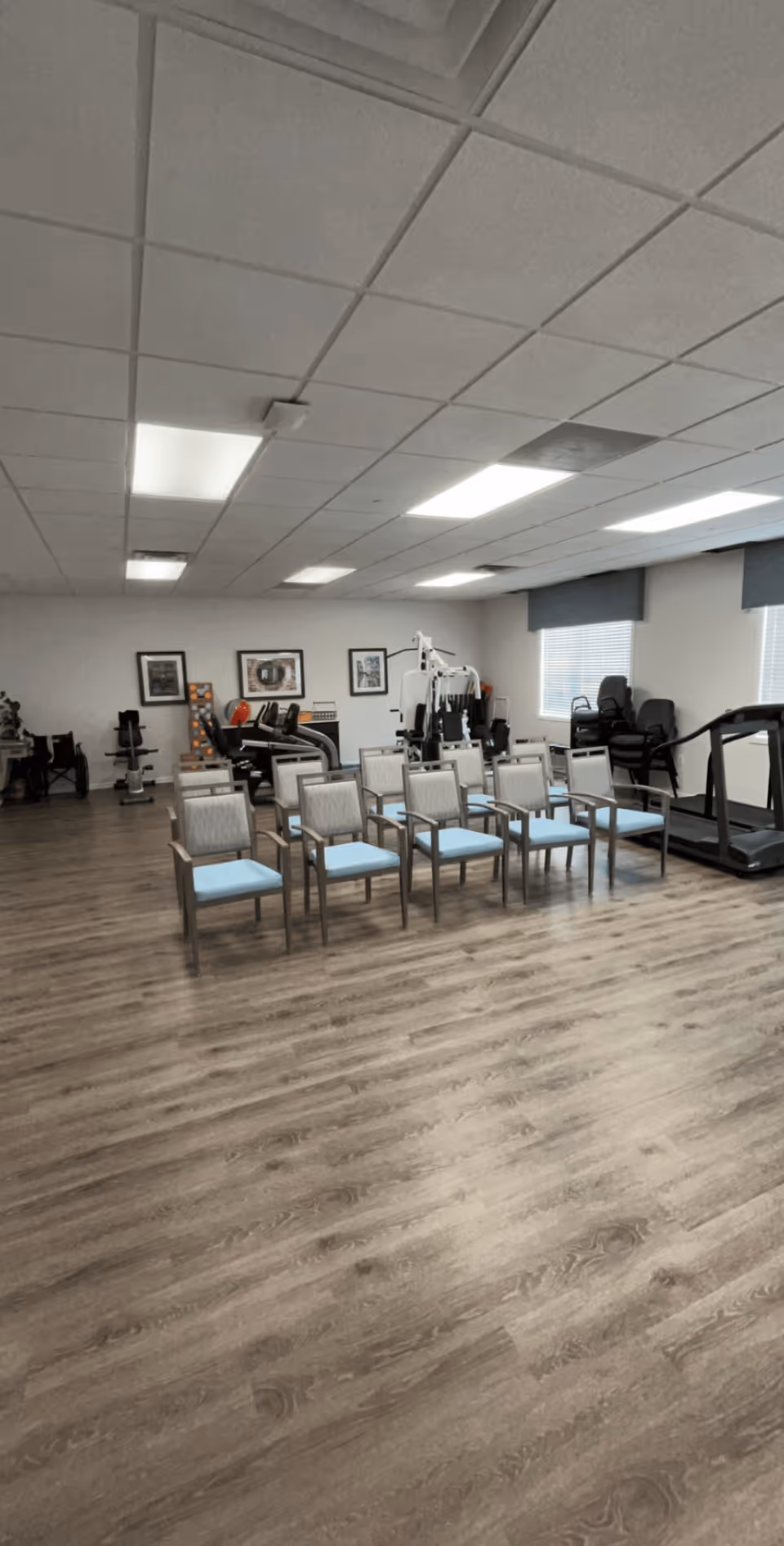 A spacious room with wooden flooring featuring a row of chairs with light blue cushions in the center. In the background, there is exercise equipment including a treadmill, weight machines, and other fitness gear. The room has white walls with framed pictures and large windows with gray blinds allowing natural light to enter.