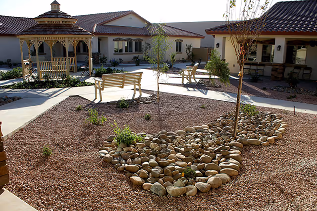 Outdoor courtyard area of Carlton Village Senior Care Community featuring a gazebo, benches, small trees, and landscaped rock beds with a building in the background under a clear sky.