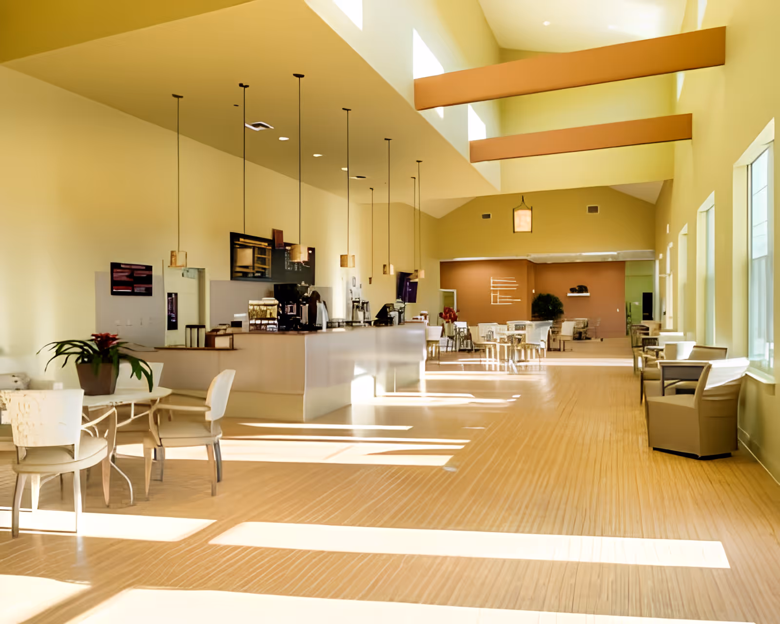 Bright and spacious interior of a retirement community dining area with high ceilings and large windows letting in natural light. The room features a long counter with hanging pendant lights, several tables and chairs arranged for dining or socializing, and decorative plants.