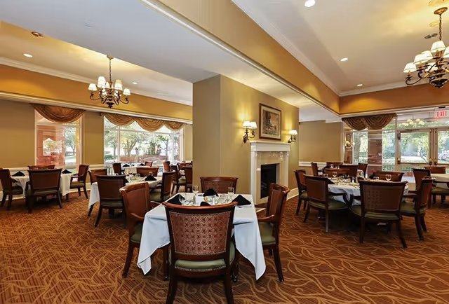 A spacious dining room with multiple tables covered in white tablecloths and set with glassware and napkins. The room features large windows with brown valances, a carpeted floor with a patterned design, chandeliers, and a fireplace with wall sconces on either side. Outside greenery is visible through the windows.