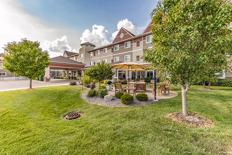 Front exterior of a senior living facility with a landscaped lawn, patio seating under umbrellas, and a covered entrance.
